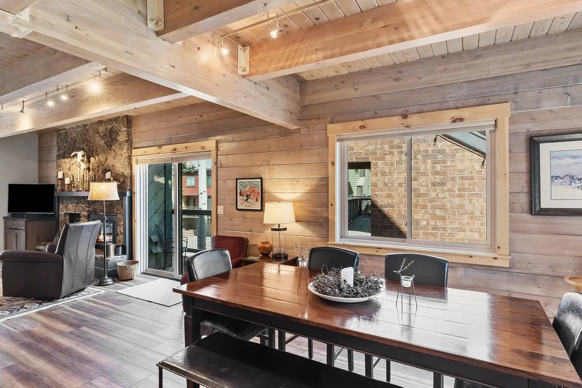 A warm, inviting dining area featuring a large table, a glowing fireplace, and expansive windows. The scene is set for family dinners, socializing by the fire, or sharing snacks after skiing.