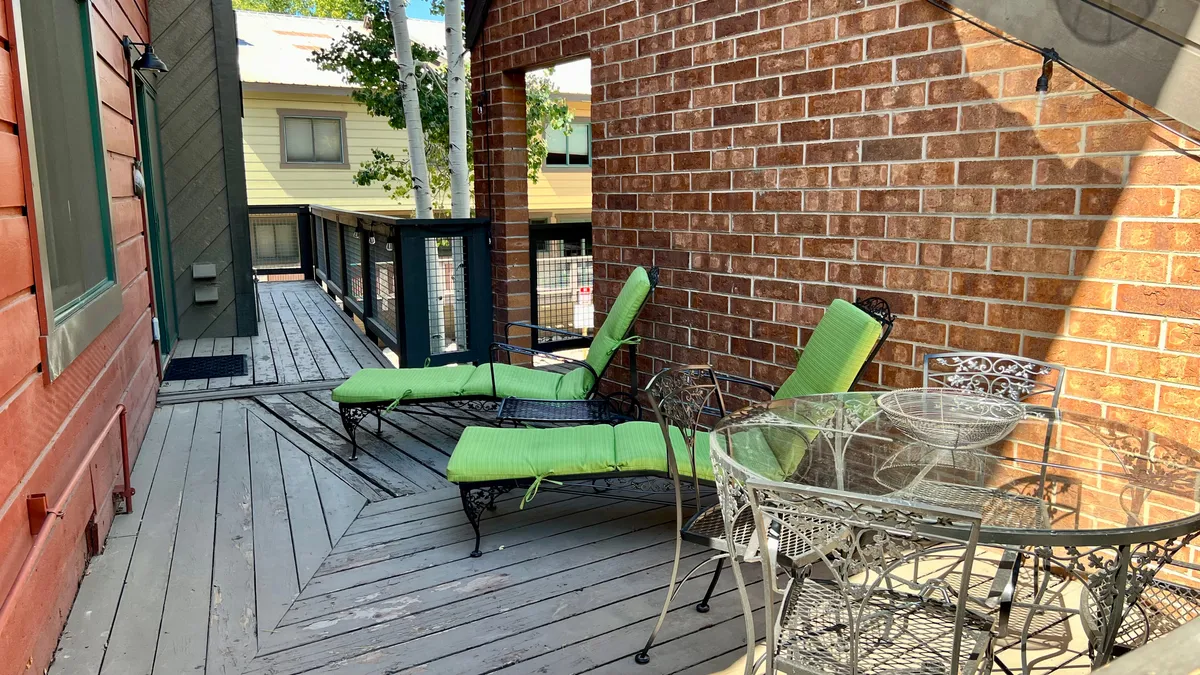 This exterior image shows green lounge chairs and a glass table nestled on a patio alongside a brick wall. The scene suggests a welcoming and communal outdoor space ideal for relaxation, sunbathing, socializing, or enjoying a meal. The casual setup hints at the community's inviting atmosphere and offers a glimpse of resort-style perks beyond the front door.