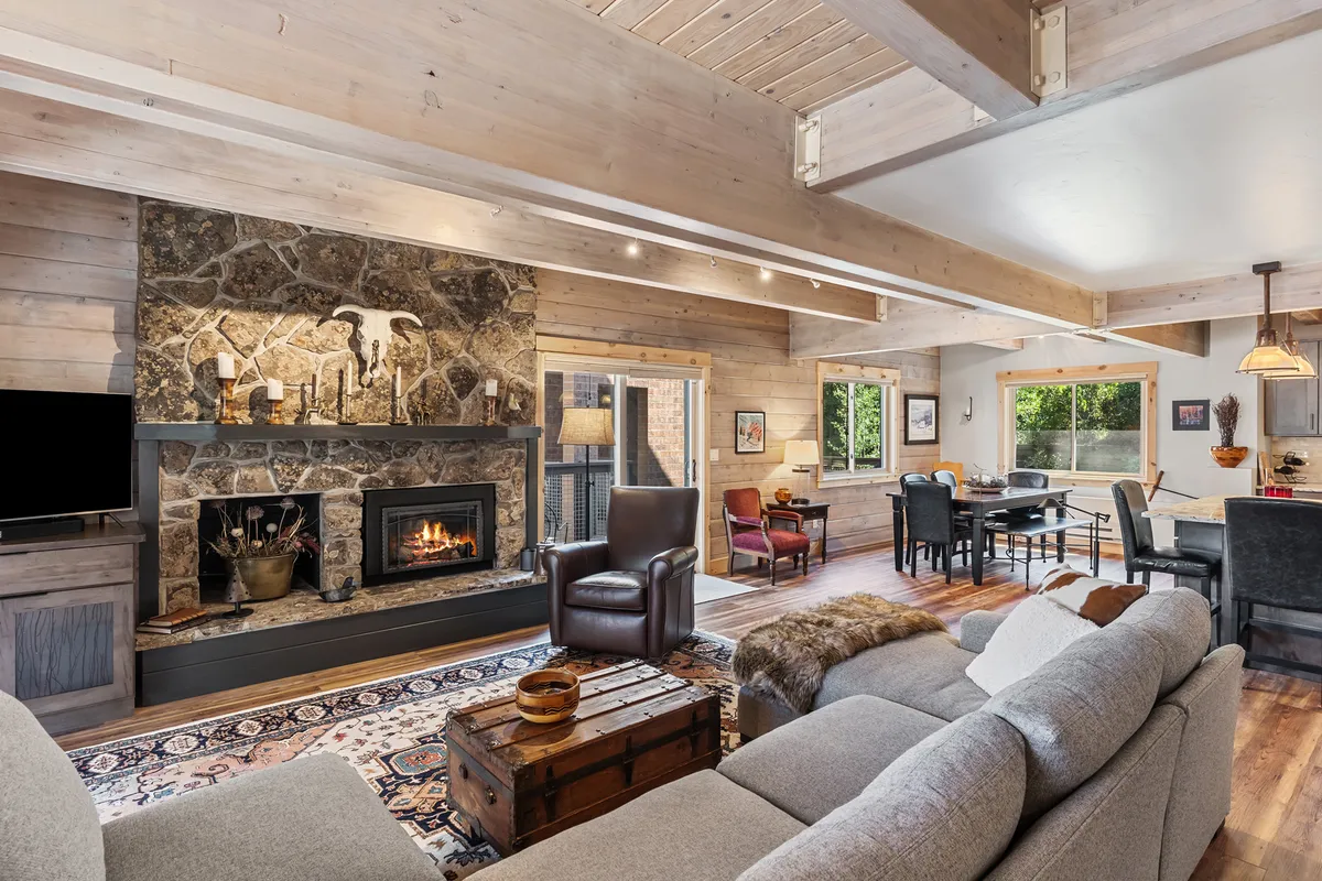 The image showcases a warm, modern living and dining area featuring a crackling fireplace, plush sofa, wooden beams overhead, large windows that welcome in mountain light, and an inviting dining table perfect for gathering. The ambiance is cozy, ideal for relaxing, sharing meals, and spending quality time together.
