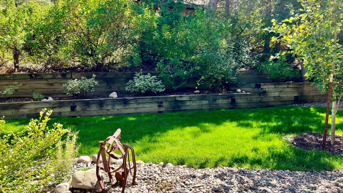 This exterior image features a green grass lawn bordered by a wooden retaining wall and accented by a rusted metal sculpture. The space is sunlit and inviting, suggesting a peaceful outdoor environment suitable for relaxation and informal gatherings.