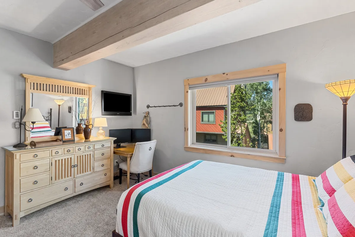 A tranquil bedroom with a striped blanket on the bed, a small desk and chair for work or play, a dresser with a mirror, and a view of trees outside the window. The room is naturally lit and inviting.