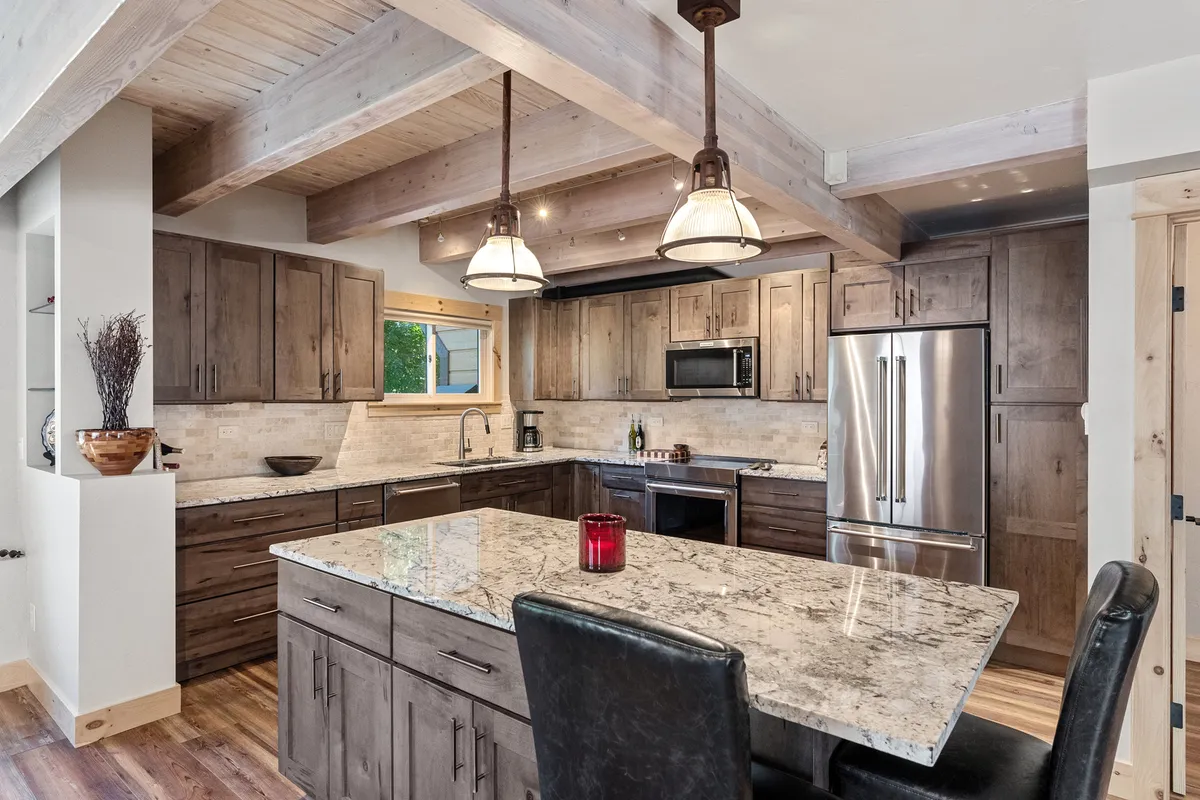 A close-up of the modern gourmet kitchen, featuring a sleek granite island, stainless steel appliances, and exposed wooden beams above. The space exudes both functionality and warmth, perfect for cooking, entertaining, or starting the day with breakfast at the bar.