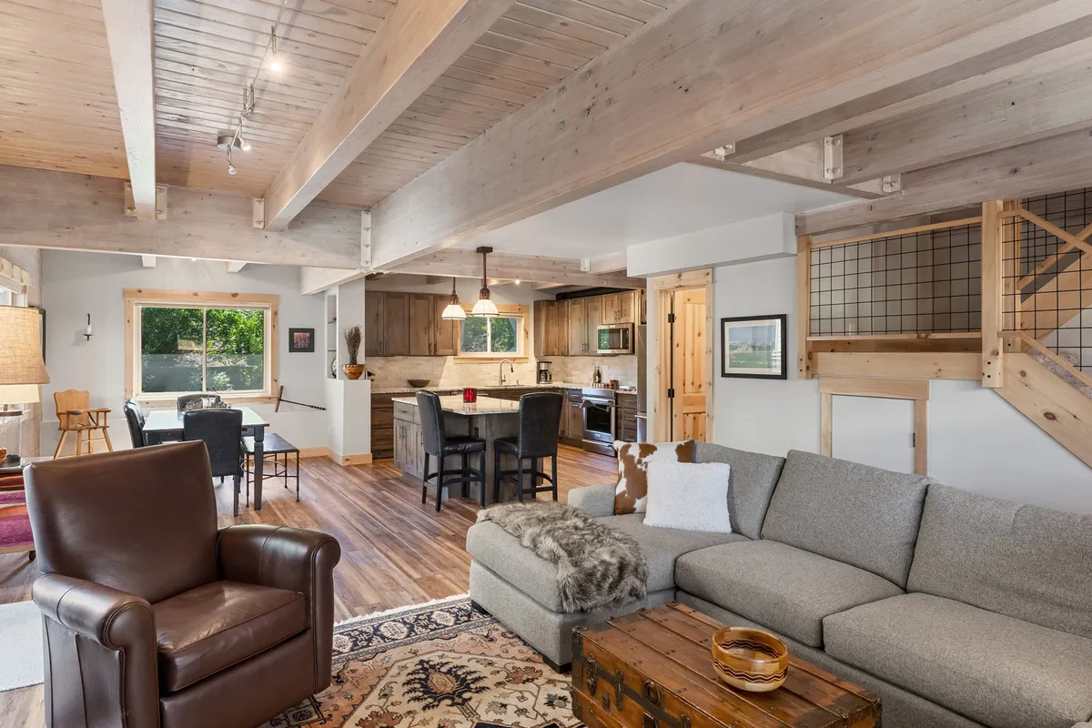 This image beautifully captures the open-concept main living area, seamlessly connecting the living room, dining table, and kitchen island under warm wooden beams. The scene features a comfortable sofa, a spacious dining table perfect for group gatherings, and a modern kitchen with an inviting island, all bathed in soft natural light.