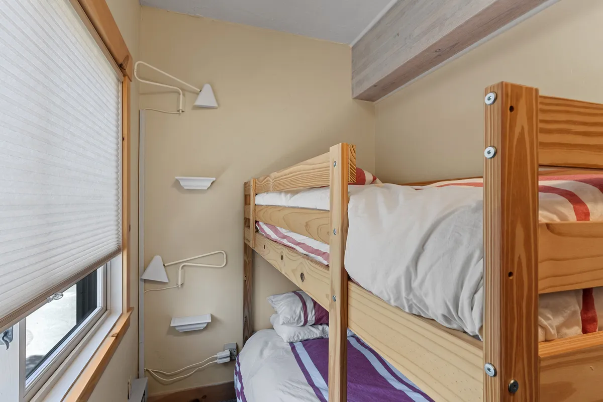 A cozy bunk room with built-in bunk beds, wall-mounted reading lights, and a window allowing daylight in. The space is designed for both relaxation and fun, suitable for children or extra guests.