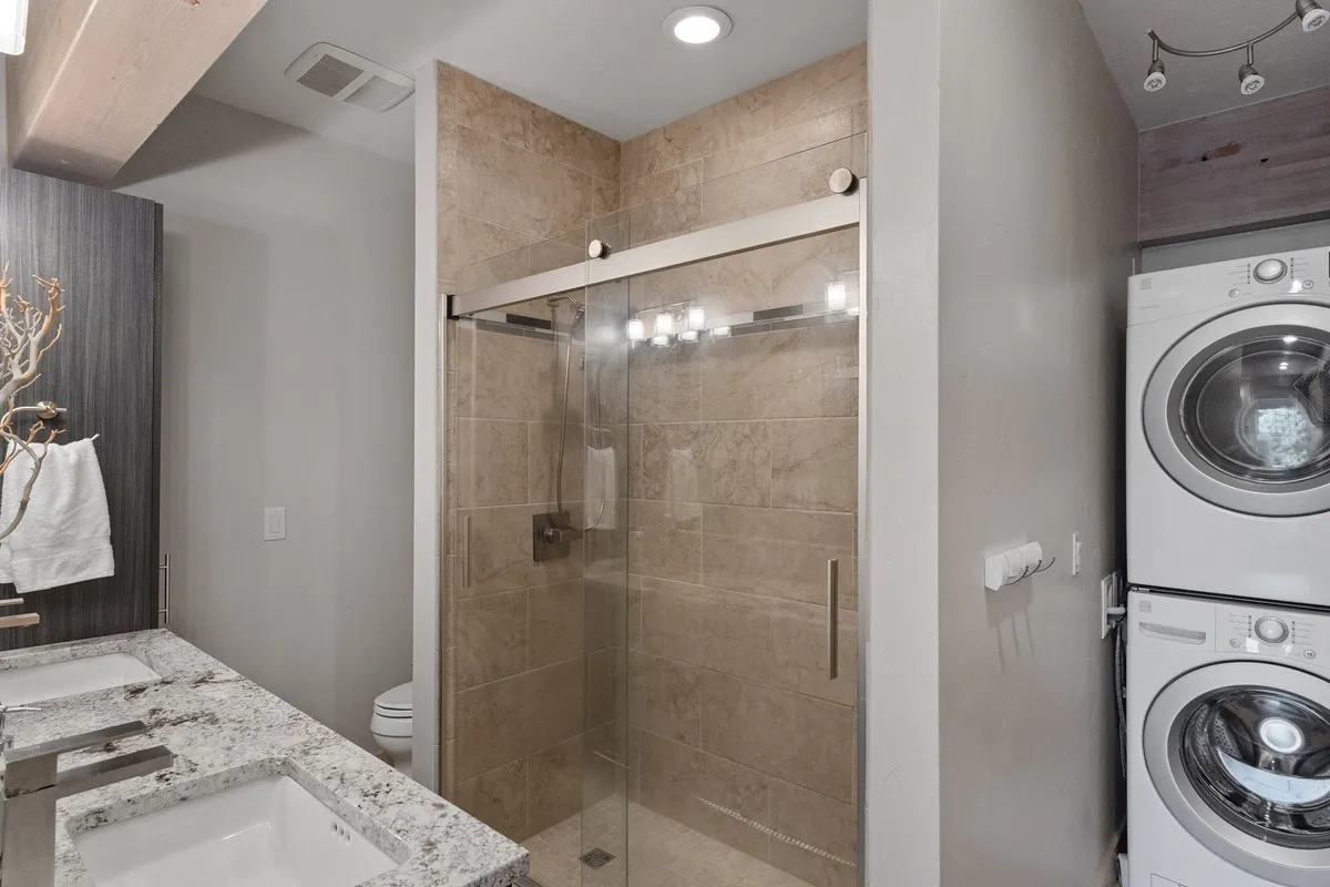A modern bathroom with a glass shower enclosure, double granite sinks, and a stacked washer and dryer. The clean lines and practical amenities emphasize comfort and convenience, echoing the spa-inspired vibe.