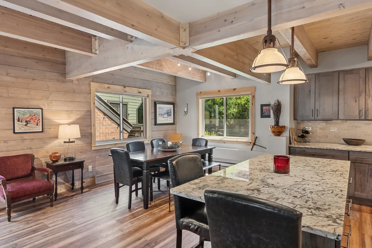 A bright, open dining space with a granite countertop, dining table, large window for natural light, and exposed wooden beams. The room feels cozy and is well-suited for family meals or relaxing after an active day.
