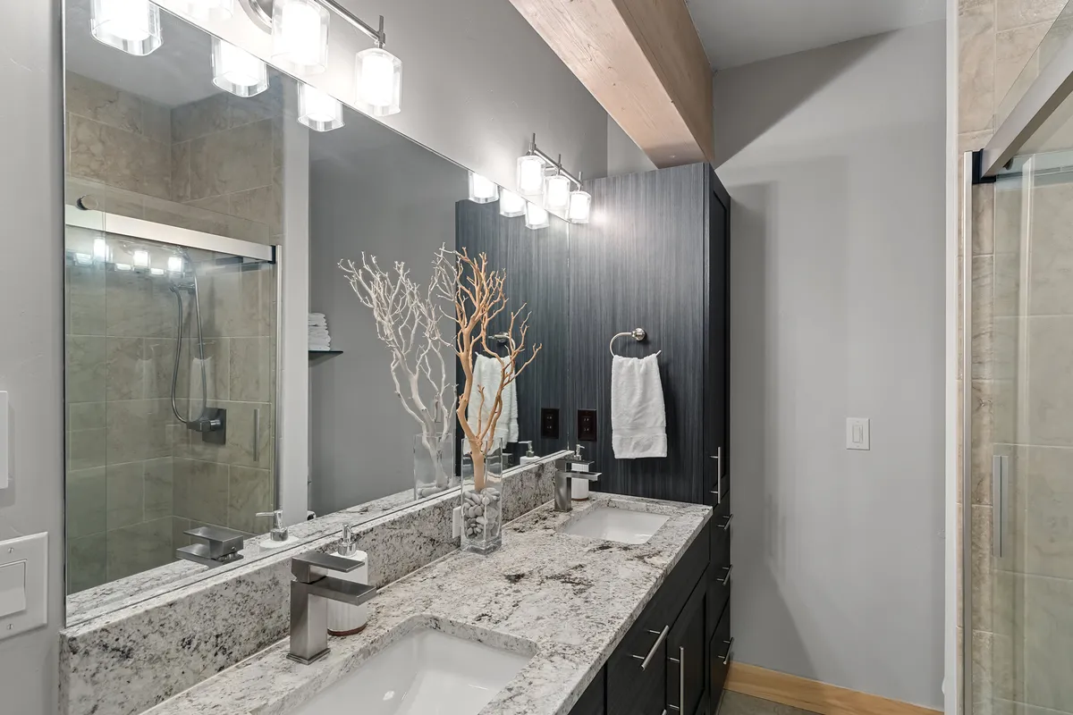 A bright, spa-inspired bathroom with double vanity sinks, large mirrors reflecting morning sunlight, a decorative branch centerpiece, and a spacious shower enclosure. The atmosphere is modern and calm, inviting self-care and rejuvenation.