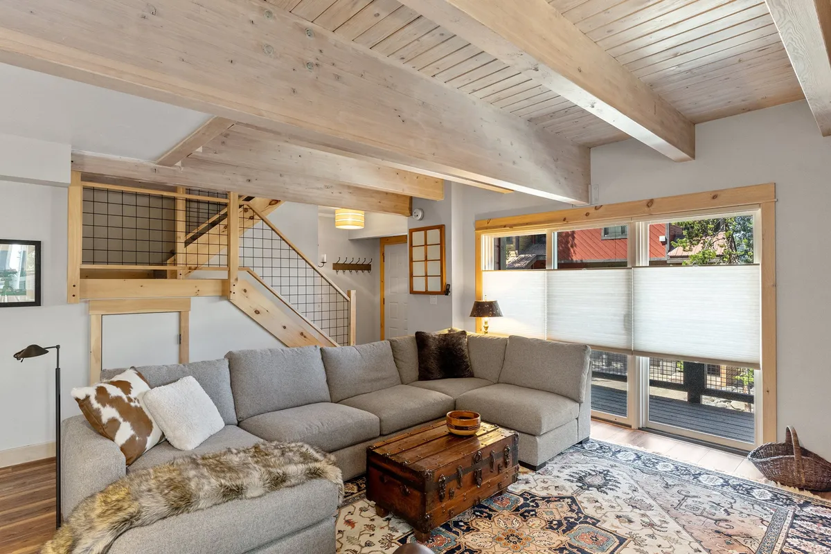 A spacious living area with a large sectional sofa, wooden beam ceiling, and a vintage trunk coffee table. Sunlight spills across the area rug, highlighting a space ideal for unwinding, playing games, or gathering together after a day outdoors.