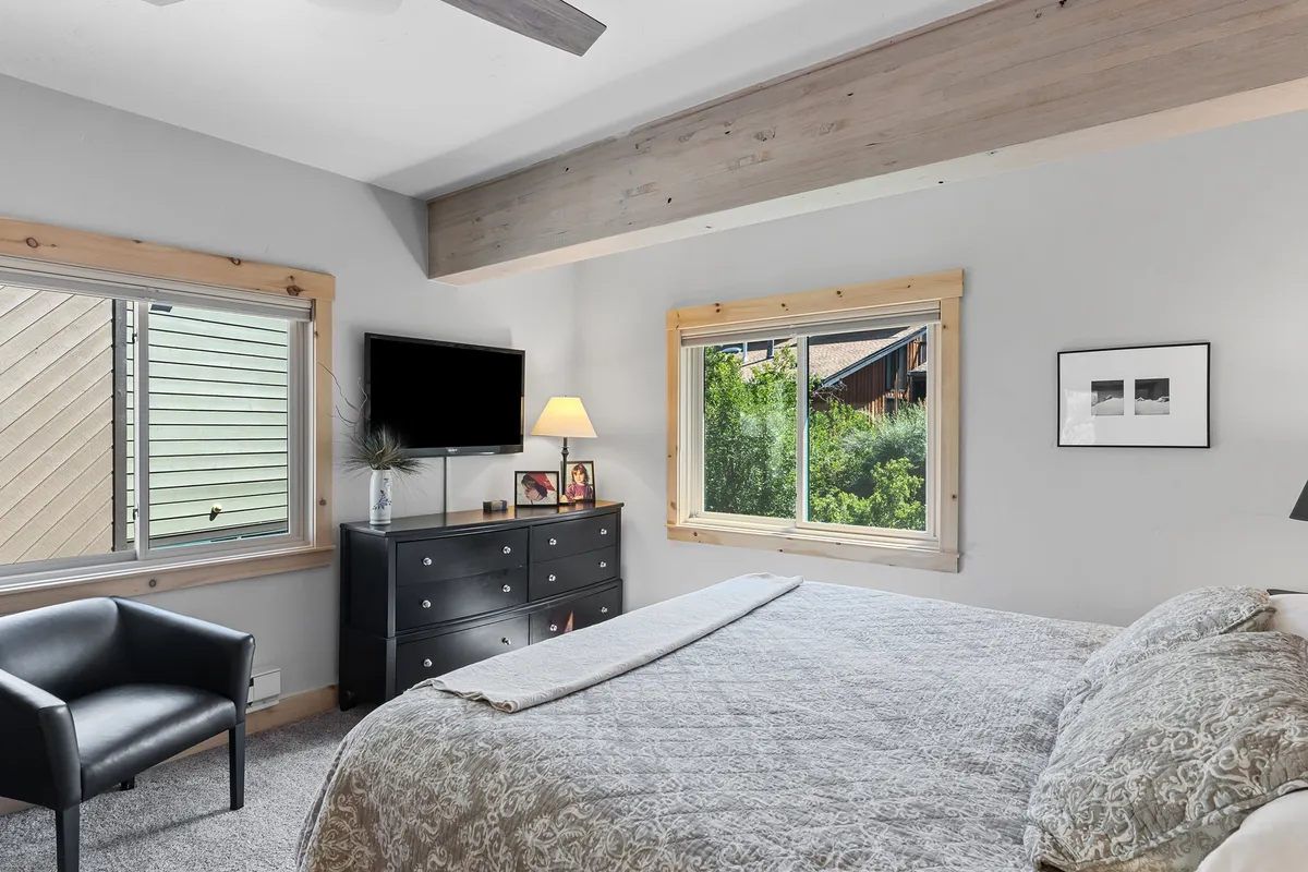 A serene bedroom with a large bed, soft natural light pouring in through the windows, a comfortable armchair, and warm ambient lighting from a lamp. The space feels airy and private, providing a restful retreat with views that hint at the mountain setting.