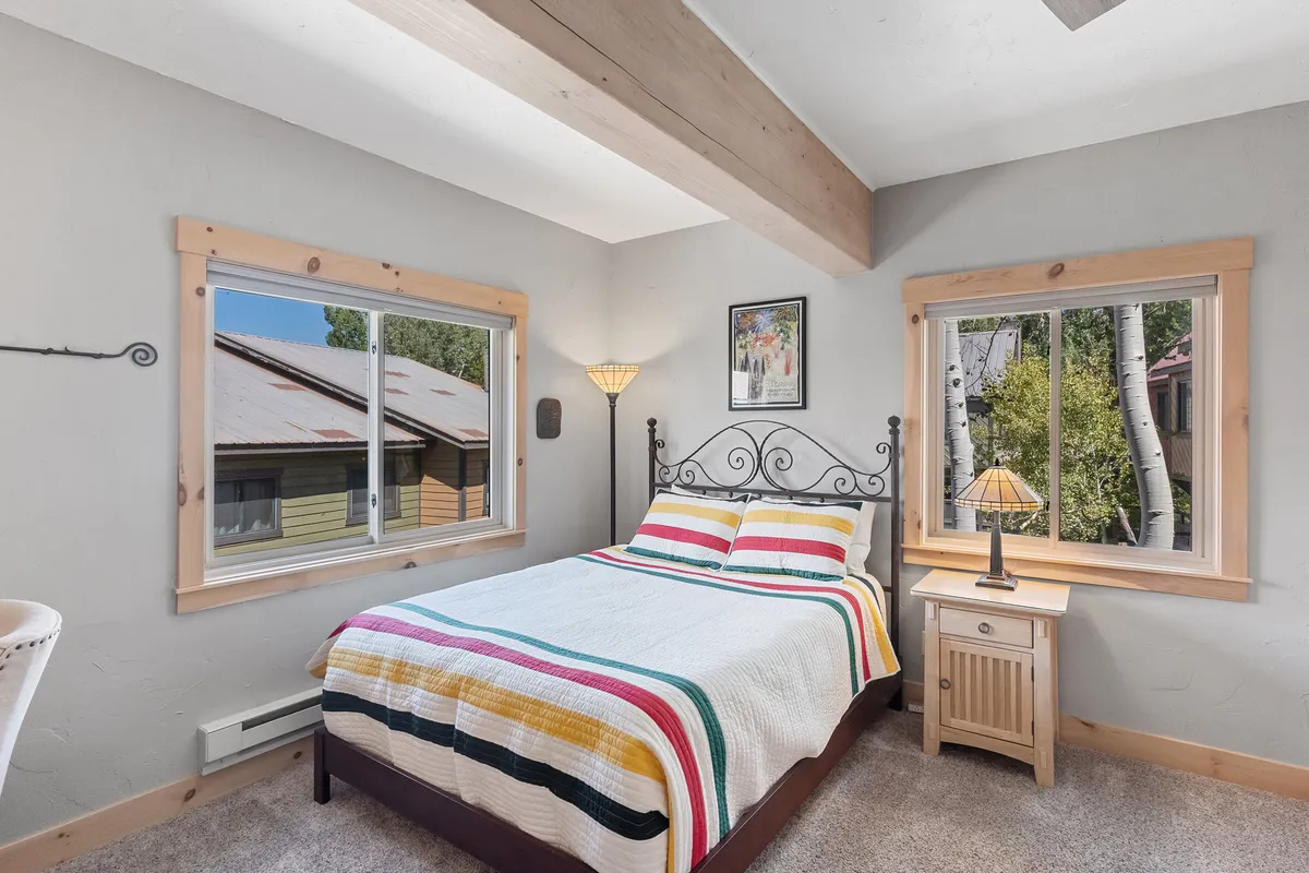A cozy bedroom with a bed covered in a colorful quilt, large windows that fill the space with natural light, and a decorative lamp for evening relaxation. Framed artwork adds a personal touch.